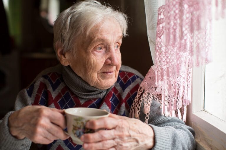 Elderly woman drinking tea sitting near the window.
