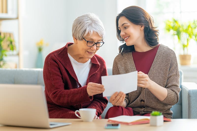 Two women, sitting on the sofa with a paper receipt, are calculating expenses, managing the family budget.