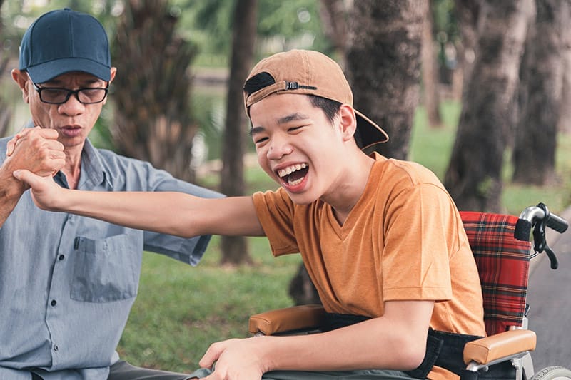 Asian happy disabled teenage boy on wheelchair smiling face with arm muscle strength test with parent, Male style play in teen and lifestyle of diverse family.