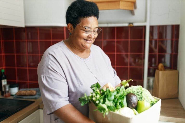 African American senior woman holding big box with green fresh vegetables standing in her kitchen