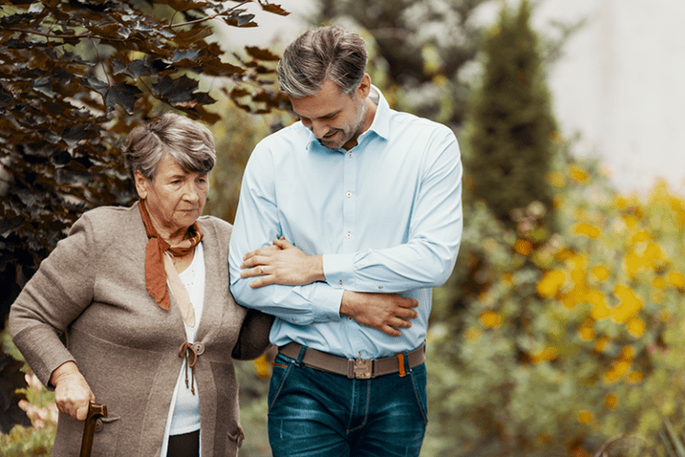 Older woman walking with a cane through a garden with her son or caregiver