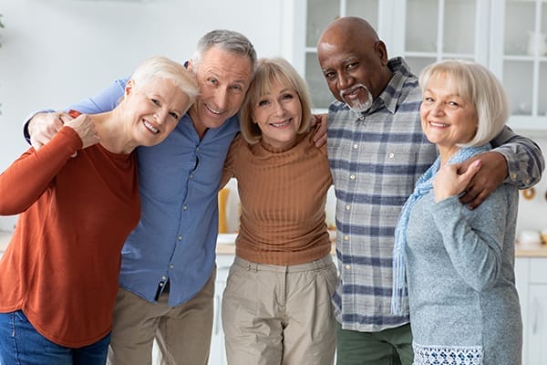Group photo of happy elderly people different nationalities standing at kitchen, hugging and smiling at camera, senior men and women having party at home, spending weekend together